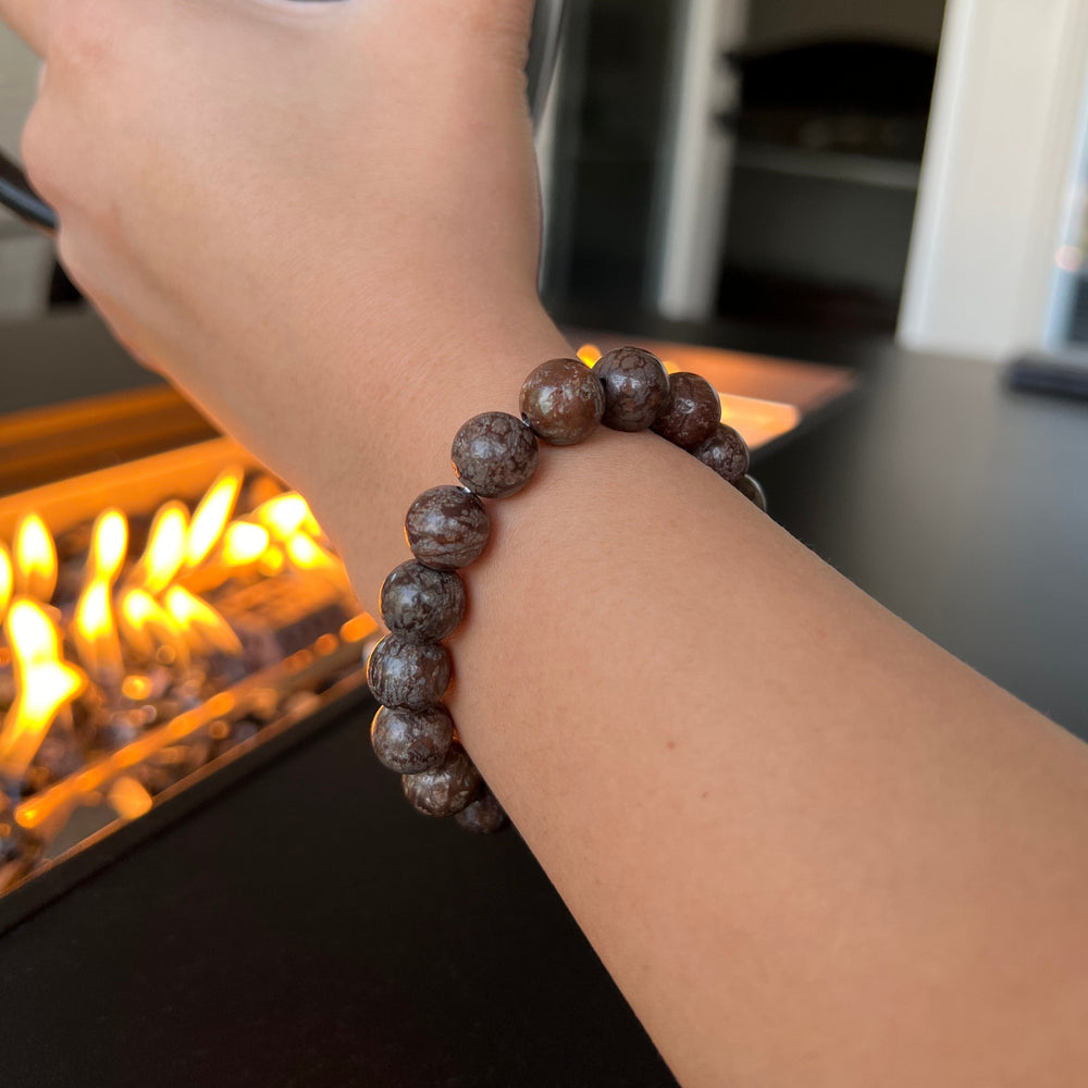 Close-up of a woman's wrist wearing a red snowflake stretch style bracelet made of 10mm polished stone beads on a clear stretch cord, with a blurred lit firepit in the background.