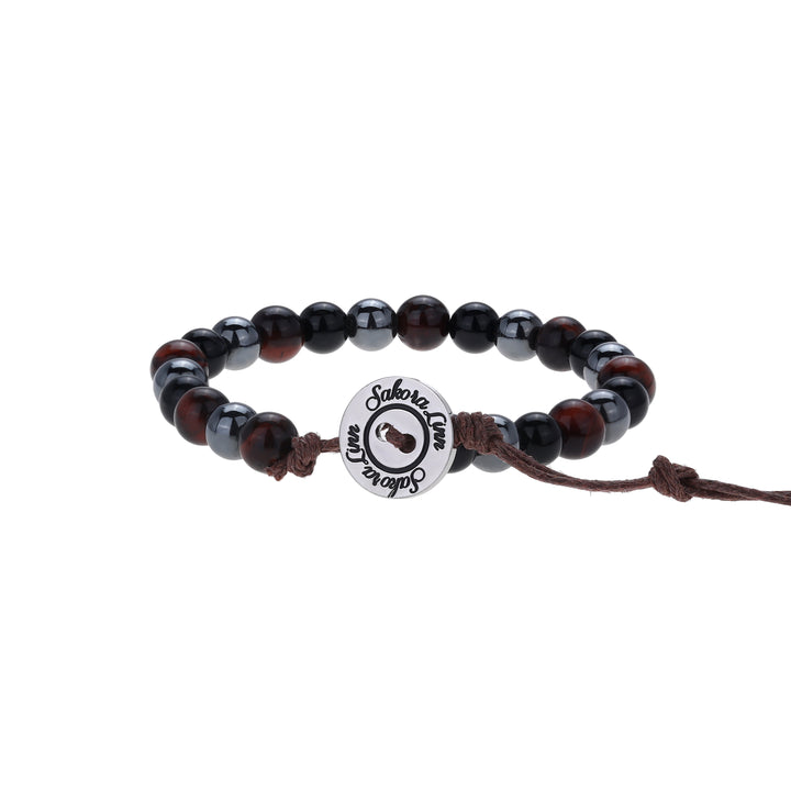 Close-up of a bead bracelet made with 6mm tiger's eye, black onyx, and hematite beads, in black, brown and dark gray colors, strung on an adjustable brown cord, with a silver color logo button closure stamped in black wth Sakora Linn, displayed on a white background.