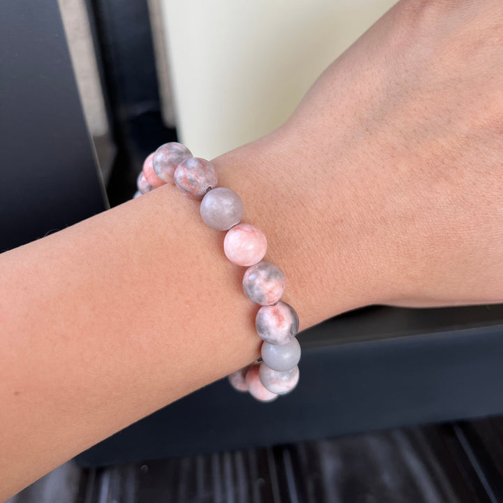 Close-up of a woman's wrist wearing a rhodonite stretch style bracelet made with 10mm round rhodonite beads in shades of pink and gray, with a gray and white blurred background.