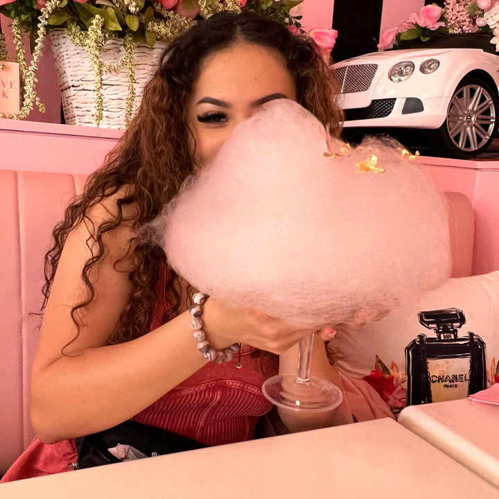 A woman with long curly brown in a pink dress, sitting in a pink booth holding pink cotton candy, wearing  pink and gray 12mm rhodonite bracelet.