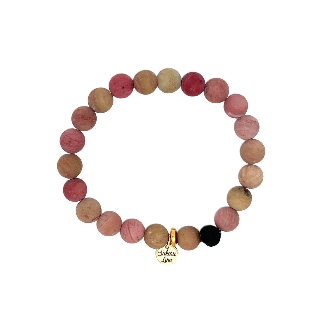 Close-up of a stretch style rhodonite bracelet made with 8mm round matte pink rhodonite beads, in shades of pink and tan, with one black clay bead, an 18k gold plated logo tag engraved with Sakora Linn, displayed on a white 
background.