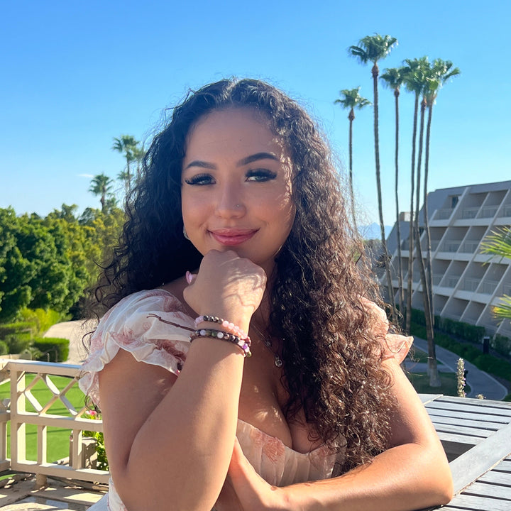 Close-up of a woman sitting on a balcony, with long black curly hair wearing a white and pink print dress with her hand resting on her face, wearing two beaded bracelets on her wrist, in shades of pink, green, purple, and black including the rose quartz amethyst rhodonite wrap bracelet, with a gray building, green grass and green trees in the background.