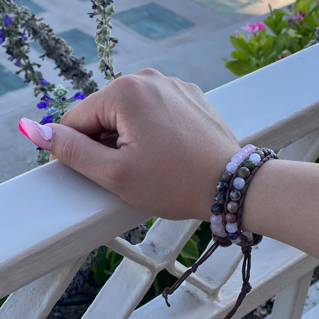 Close-up of a woman's hands with long pink fingernails, resting on a white balcony railing, wearing two beaded bracelets on her wrist, in shades of pink, green, purple, and black including the rose quartz amethyst rhodonite wrap bracelet, with a blurred sidewalk and green, pink, and purple flower plants in the background.