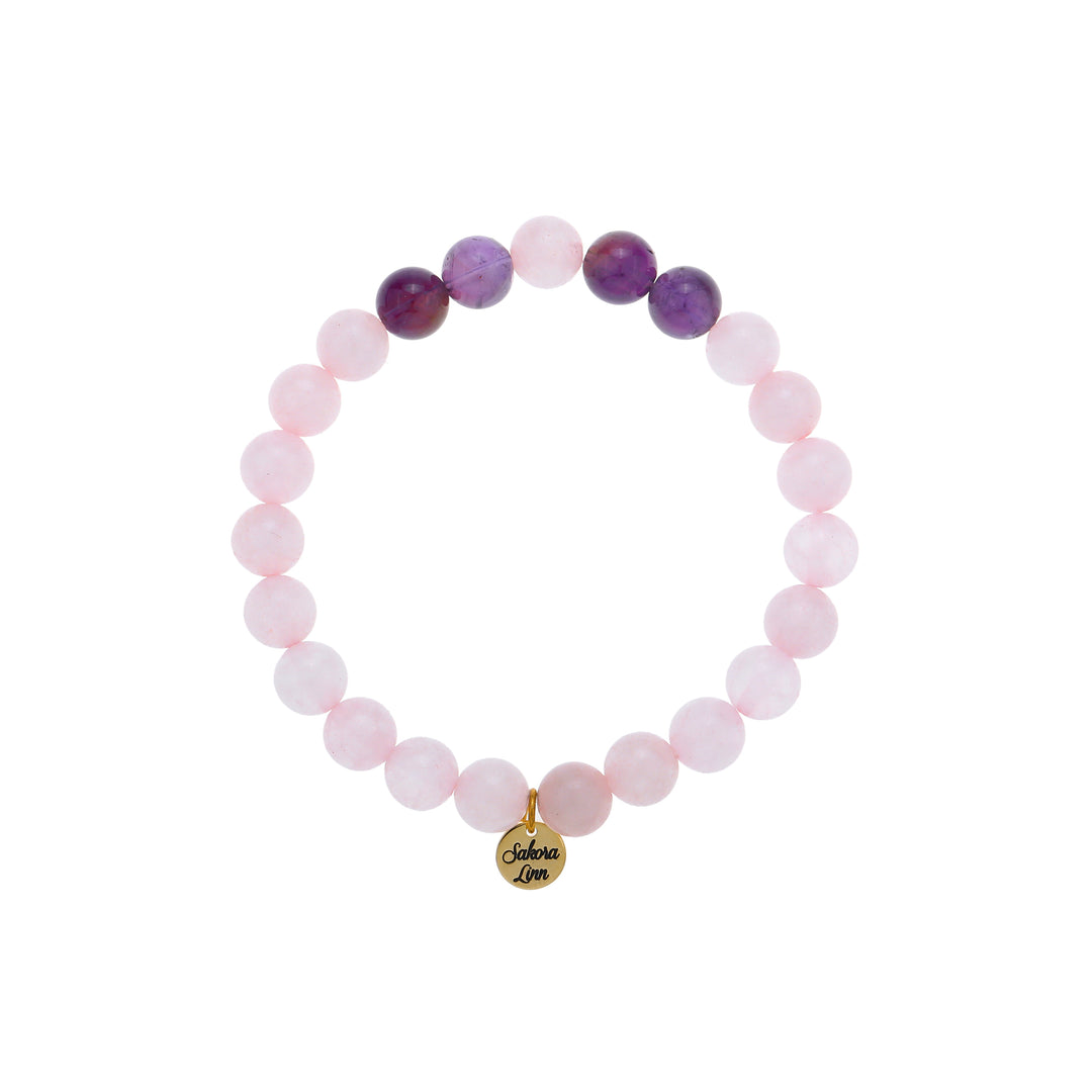 Close-up of a stretch style rose quartz bracelet made with 8mm round rose quartz beads with four 8mm amethyst beads on each side of one pink center bead, in shades of pink and purple, with an 18k gold plated logo tag engraved with Sakora Linn, displayed on a white background.
