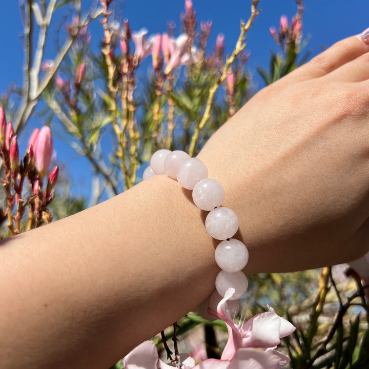 A woman's wrist wearing a pale light pink 12mm bead rose quartz bracelet, with blurred sky and pink flowers in the background.
