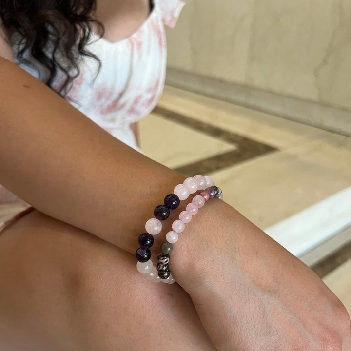 Close-up of a woman's wrist resting on her bare knee, wearing two beaded bracelets on her wrist, in shades of pink, green, purple, and black including the rose quartz rhodonite bracelet, with a blurred tan marble floor and her white and pink print dress, and long black hair, in the background.