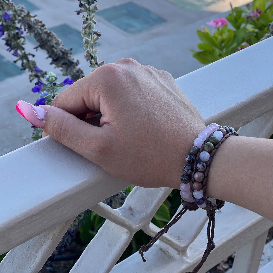 Close-up of a woman's hands with long pink fingernails, resting on a white balcony railing, wearing two beaded bracelets on her wrist, in shades of pink, green, purple, and black including the rose quartz rhodonite bracelet, with a blurred sidewalk and green, pink, and purple flower plants in the background.