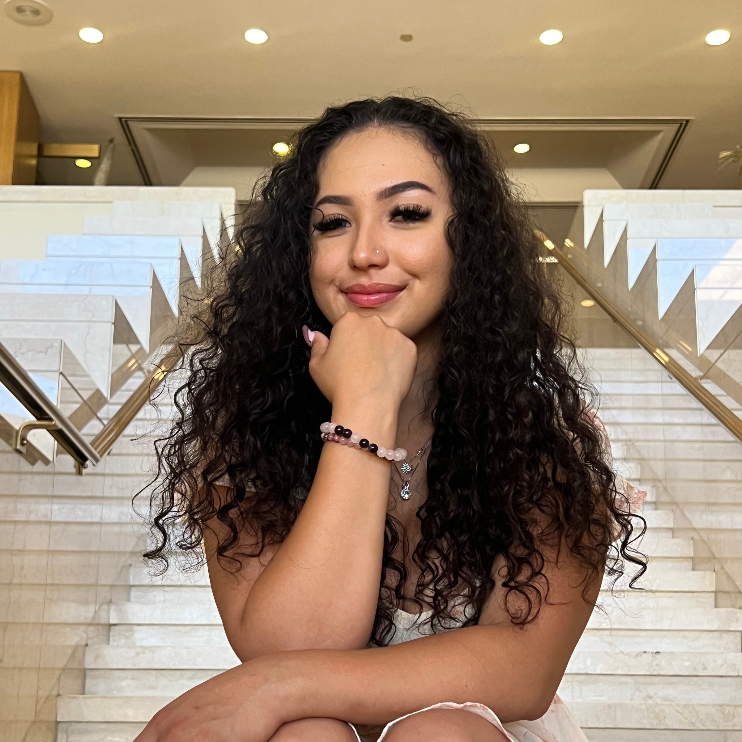 Close-up of a woman sitting on tan marble stairs with elbow resting on her leg and hand under chin, wearing two bracelets, one is a stretch style rose quartz bracelet made with 8mm round rose quartz beads with four 8mm amethyst beads on each side of one pink center bead, in shades of pink and purple, and a rose quartz and rhodonite bracelet with 6mm beads in shades of pink and black, with a marble tile background.