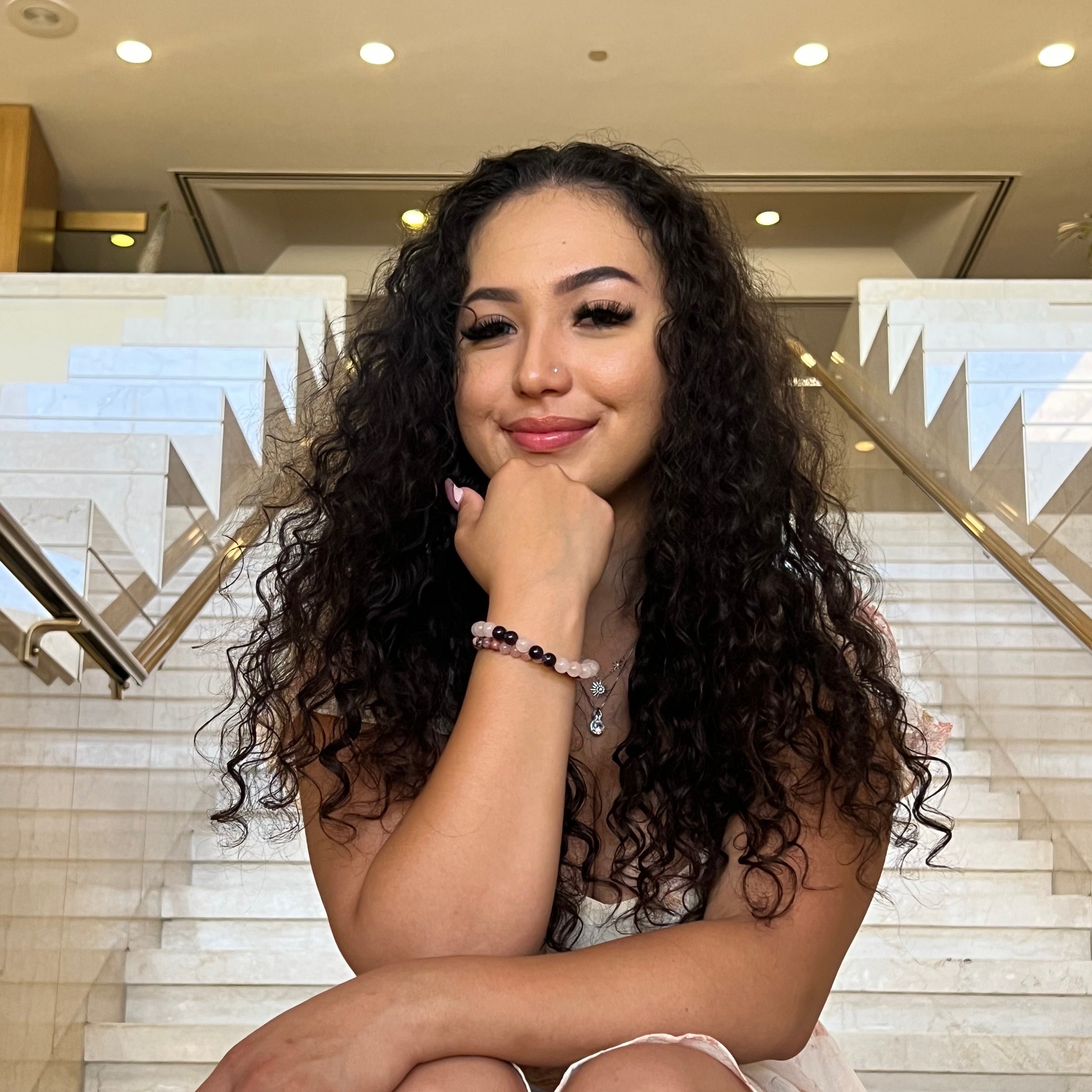 Close-up of a woman sitting on tan marble stairs with elbow resting on her leg and hand under chin, wearing two bracelets, one is a stretch style rose quartz bracelet made with 8mm round rose quartz beads with four 8mm amethyst beads on each side of one pink center bead, in shades of pink and purple, and a rose quartz and rhodonite bracelet with 6mm beads in shades of pink and black, with a marble tile background.