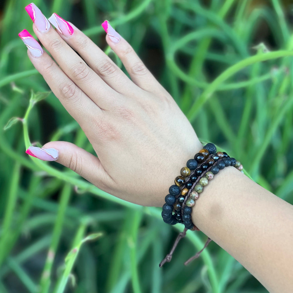 Close-up of a woman's hand with long pink fingernails, wearing three bead bracelets in shades of black, brown, and green, including the tiger's eye black onyx hematite wrap bracelet, with a blurry green plants in the background.
