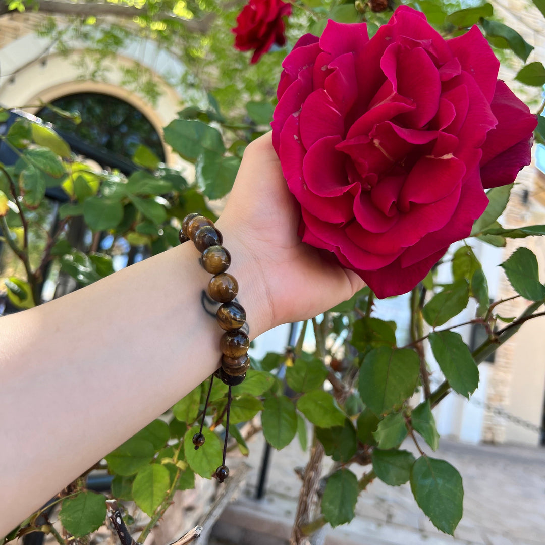 Close-up f a woman's wrist and hand, wearing a brown tiger's eye 12mm bead bracelet, holding a dark pink flower with green leaves in the background.