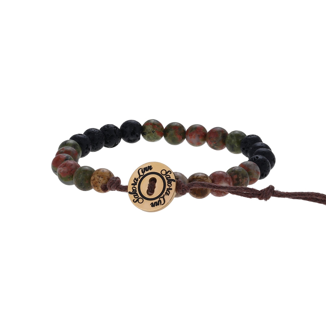 Close-up of a bead bracelet made with 6mm unakite and lava stone beads, in shades of green, black, and tan colors, strung on an adjustable brown cord, with a gold logo button closure stamped in black wth Sakora Linn, displayed on a white background.