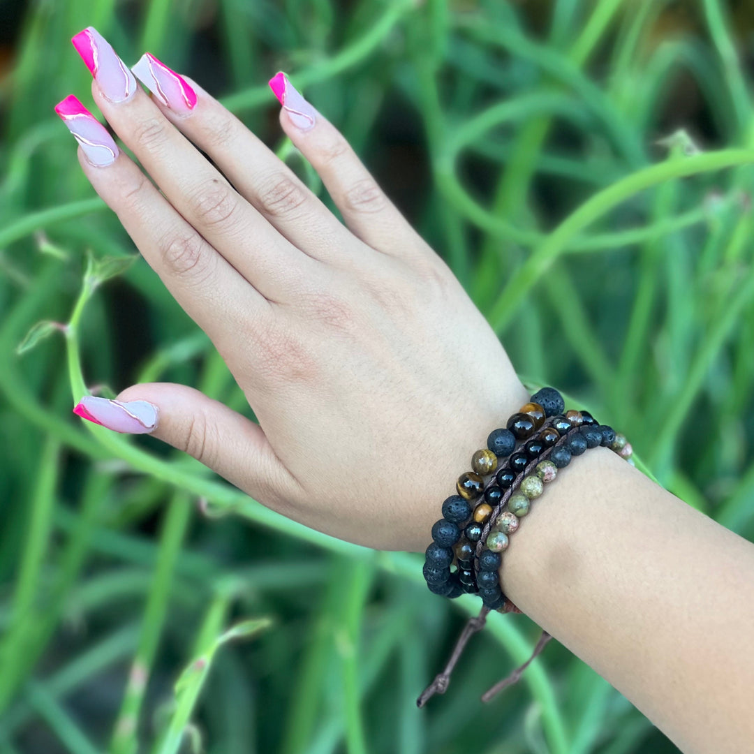 Close-up of a woman's hand with long pink fingernails, wearing three bead bracelets in shades of black, brown, and green, including the unakite lava stone bracelet, with a blurry green plants in the background.