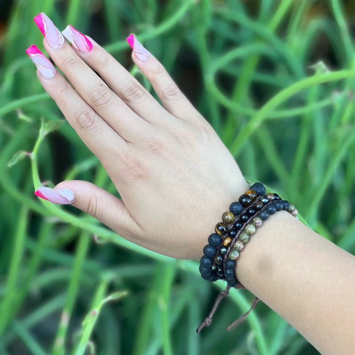Close-up of a woman's hand with long pink fingernails, wearing three bead bracelets in shades of black, brown, and green, including the unakite lava stone bracelet, with a blurry green plants in the background.