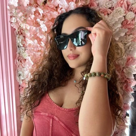 A woman with long curly brown hair in a pink top with black sunglasses wearing a green and pink unakite 12mm bead braclet, with a pink and white flower background.