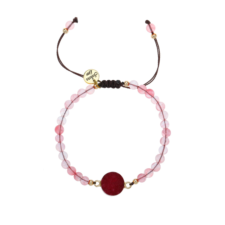 Close-up of an adjustable slip knot style bracelet with 4mm round pink beads, a 10mm deep pink red druzy stone, mounted in a gold setting, in the center of the bracelet, with small gold accent, strung on a dark brown cord with a round gold logo charm stamed with Sakora Linn in black, displayed on a white background.