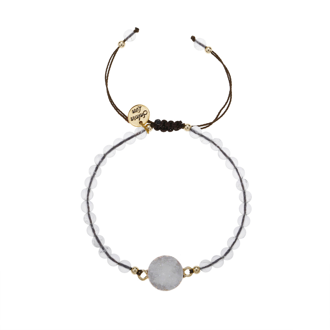 Close-up of an adjustable slip knot style bracelet with 4mm round clear white beads, a 10mm white crystal druzy stone, mounted in a gold setting, in the center of the bracelet, with small gold accent, strung on a dark brown cord with a round gold logo charm stamed with Sakora Linn in black, displayed on a white background.
