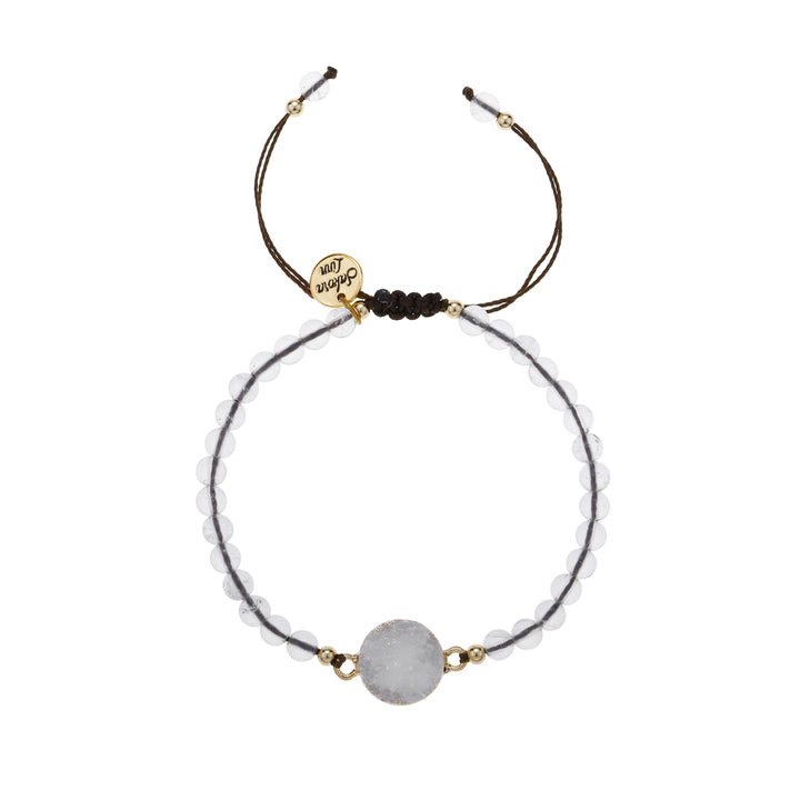 Close-up of an adjustable slip knot style bracelet with 4mm round clear white beads, a 10mm white crystal druzy stone, mounted in a gold setting, in the center of the bracelet, with small gold accent, strung on a dark brown cord with a round gold logo charm stamed with Sakora Linn in black, displayed on a white background.