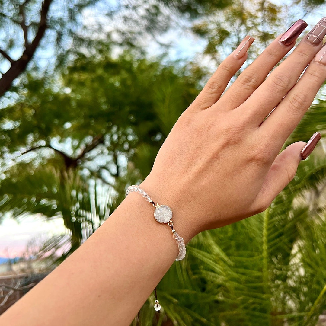 Close-up of a woman's hand with long taupe and mauve fingernails, wearing a bracelet made with clear beads and a white crystal druzy stone bead in the center, with blurred green palm trees and blue sky in the background.