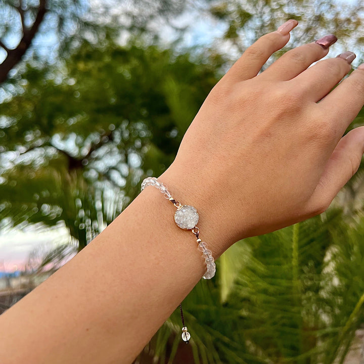 Close-up of a woman's hand with long taupe and mauve fingernails, wearing a bracelet made with clear beads and a white crystal druzy stone bead in the center, with blurred green palm trees and blue sky in the background.