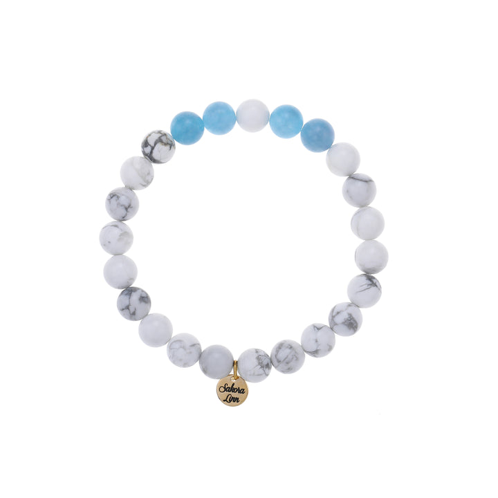 Close-up of a stretch style white turquoise and blue quartz bracelet made with 8mm round imitation white turquoise beads with four 8mm blue quartz beads on each side of one white center bead, in shades of medium blue, white, and gray, with an 18k gold plated logo tag engraved with Sakora Linn, displayed on a white background.