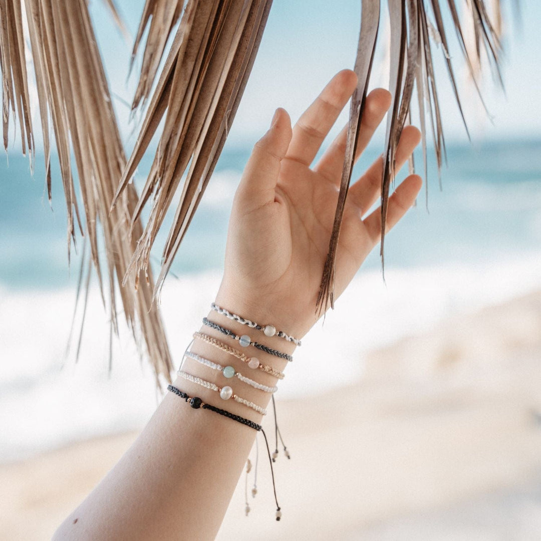 A close-up image of a young female's hand and wrist, under brwon palm tree fronds and a blurred beach sand and blue ocean in the backgraound, wearing six braided slip knot syle single stone bracelets in multiple colors, including the pearl braided bracelet.
