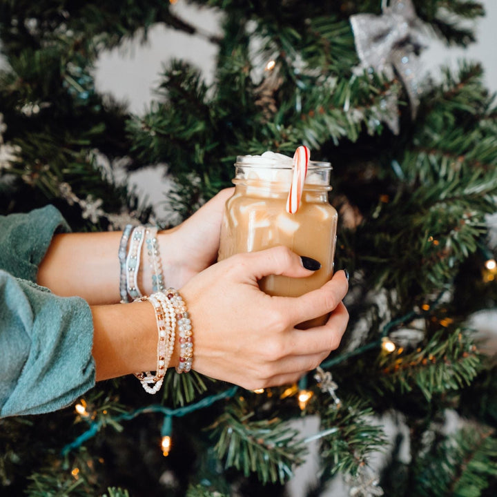 A woman's hands holding a mug of hot cocoa with candy cane and marshmellows, wearing three beaded bracelets on each wrist in shades of blue, pink, and white, with a blurred green Christmas tree in the background.