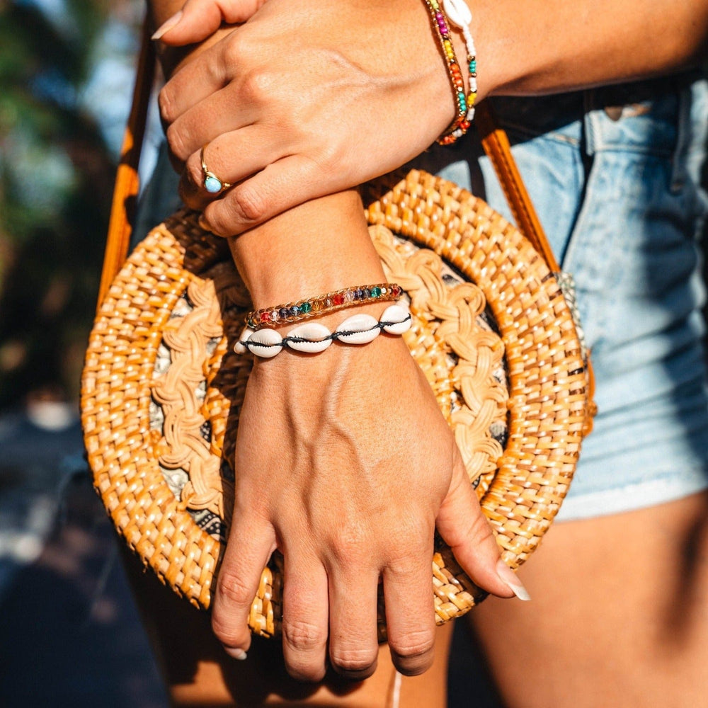 Close-up of a woman's lower torso holding a straw purse, wearing blue denim shorts and multiple bead bracelets on her wrist, including the black cowry shell bracelet.