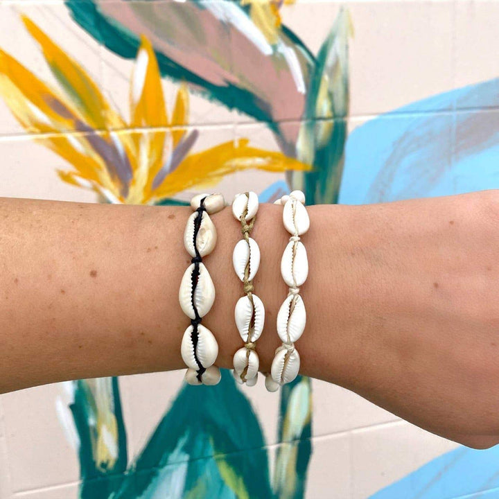 Close-up of a woman's wrist with three cowry shell bracelets, one with black cord, one with white cord, one with tan cord, with a wall painted with flowers in pastel colors.
