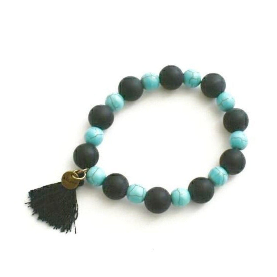 Close-up of a stretch style beaded bracelet with black onyx beads alternating with smaller turquoise magnesite beads, a black tassel, and a gold brass beljoy logo charm accent displayed on a white background.
