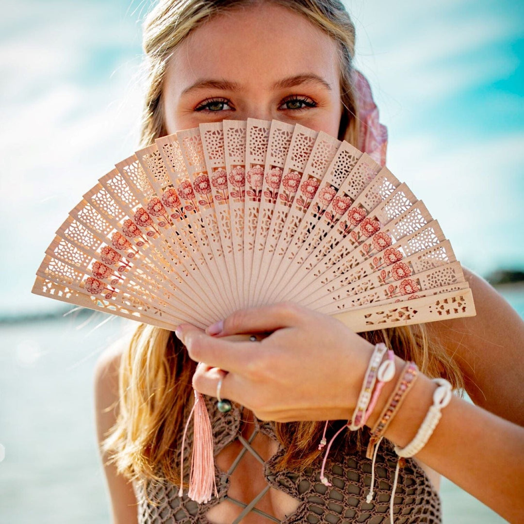 Close-up of a young woman with long blonde hair, her eyes peeking out from behind a tan oriental style fan she's holding, wearing mutiple bead bracelets on her wrist, including the blossom braided pink cowry shell bracelet.