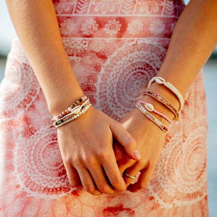 A close-up of a young woman's hands in front of her with her pink and white print flowy skirt in the background,wearing multiple beaded bracelets oh her wrists, including the pink braided blossom cowry shell bracelet.