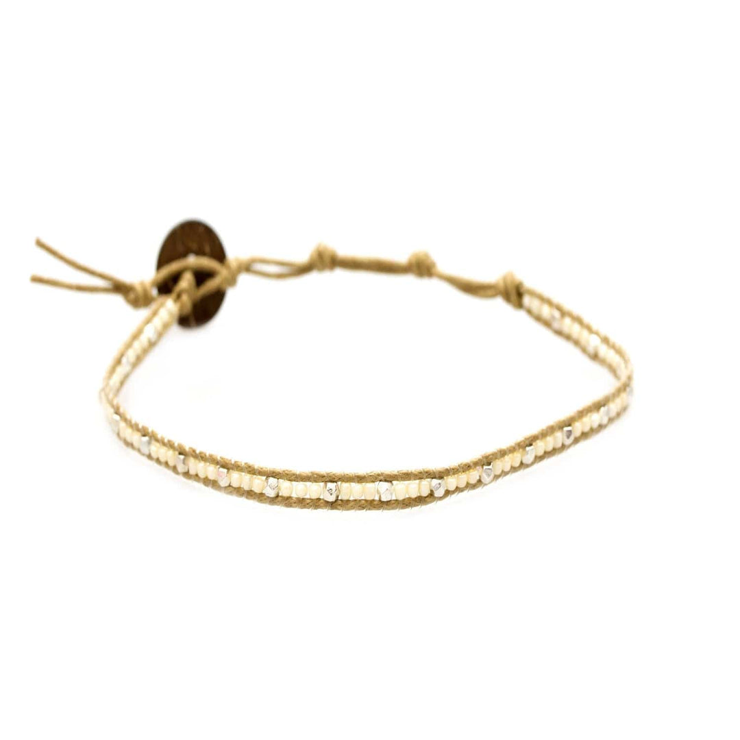 Close-up of a seed bead ankle bracelet made with pearly creamy white, and silver, seed beads, with an adustable tan colored cord and a dark brown coconut shell button closure.