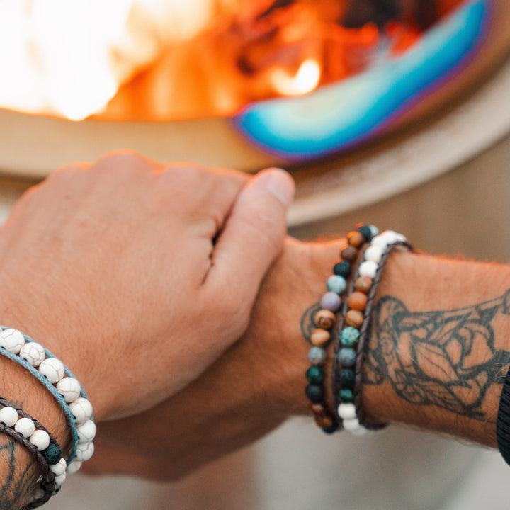 Close-up of a man's hands wearing two beaded bracelets on each wrist, with a tatoo on each wrist, with a blurred lit firepit in the background.