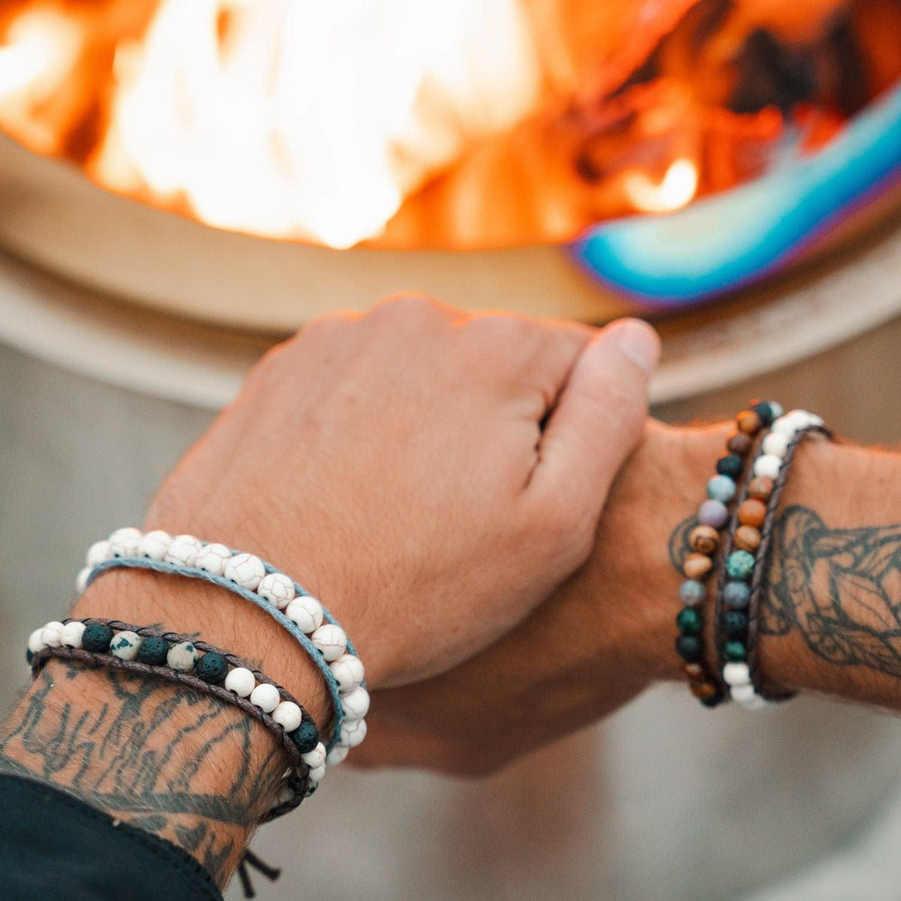 Close-up of a man's hands with a tattoo and two wrap bead bracelets on each wrist, in shades of white, black, green, and brown, with a blurred lit firepit in the background.
