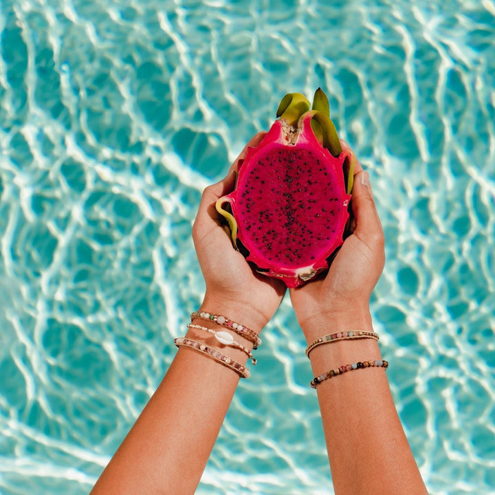 Close-up of a woman's hands holding half a pink dragonfruit, wearing three beaded bracelets on left wrist and two bead bracelets on right wrist, with blurred blue water on background.