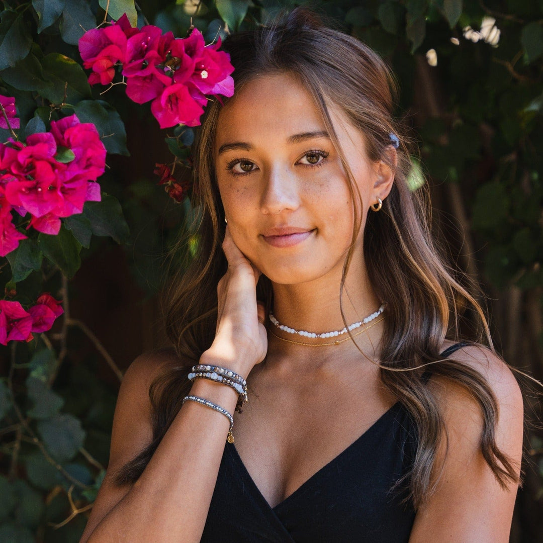 Portraint of a young woman with long brown hair and brwon eyes, wearing beaded bracelets, in shades of blue, on her wrist and a beaded choker on her neck, with a green shrub and pink flowers in background.