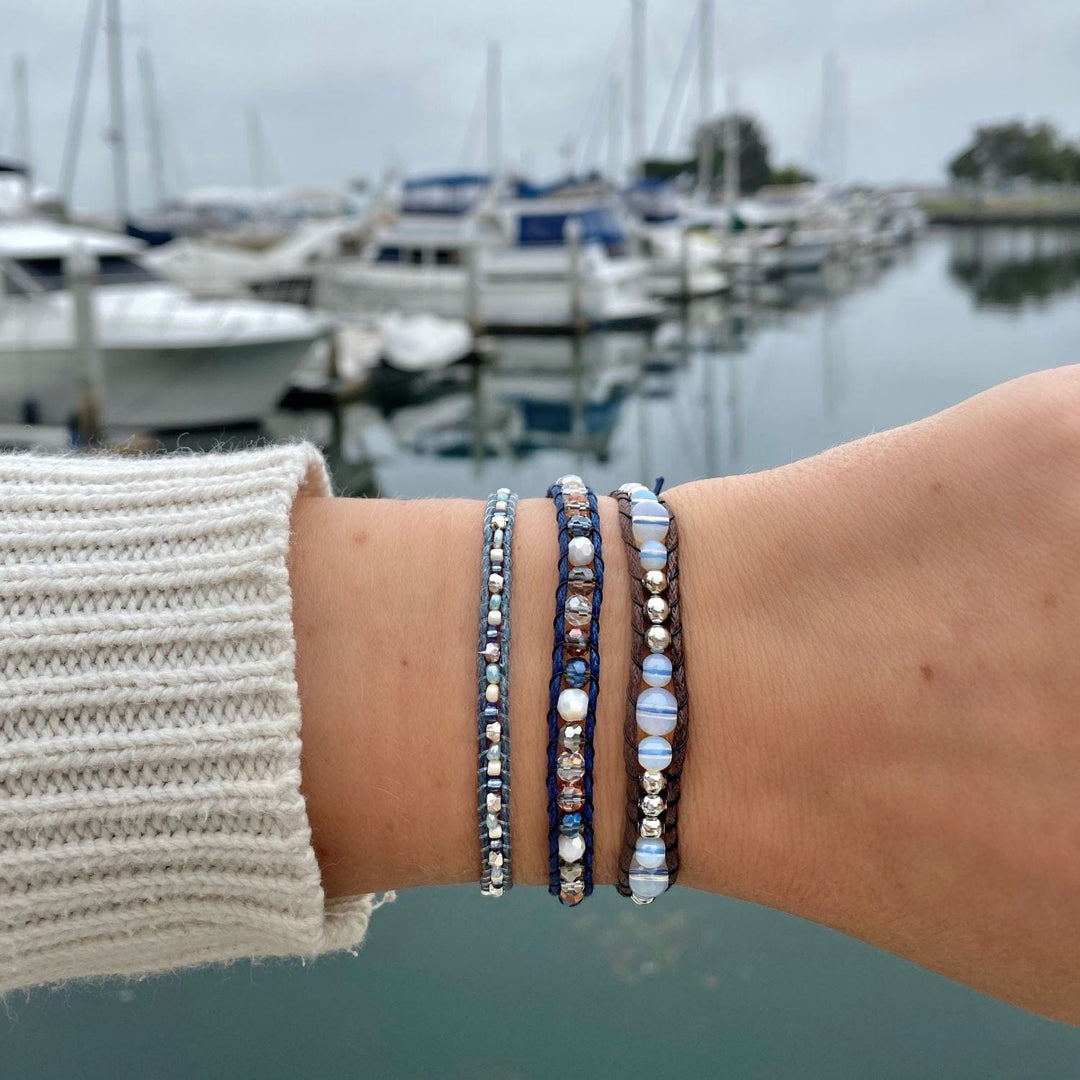 Close-up of a wrist wearing three beaded wrap style bracelets in shades of blue, with a blurred marina background.