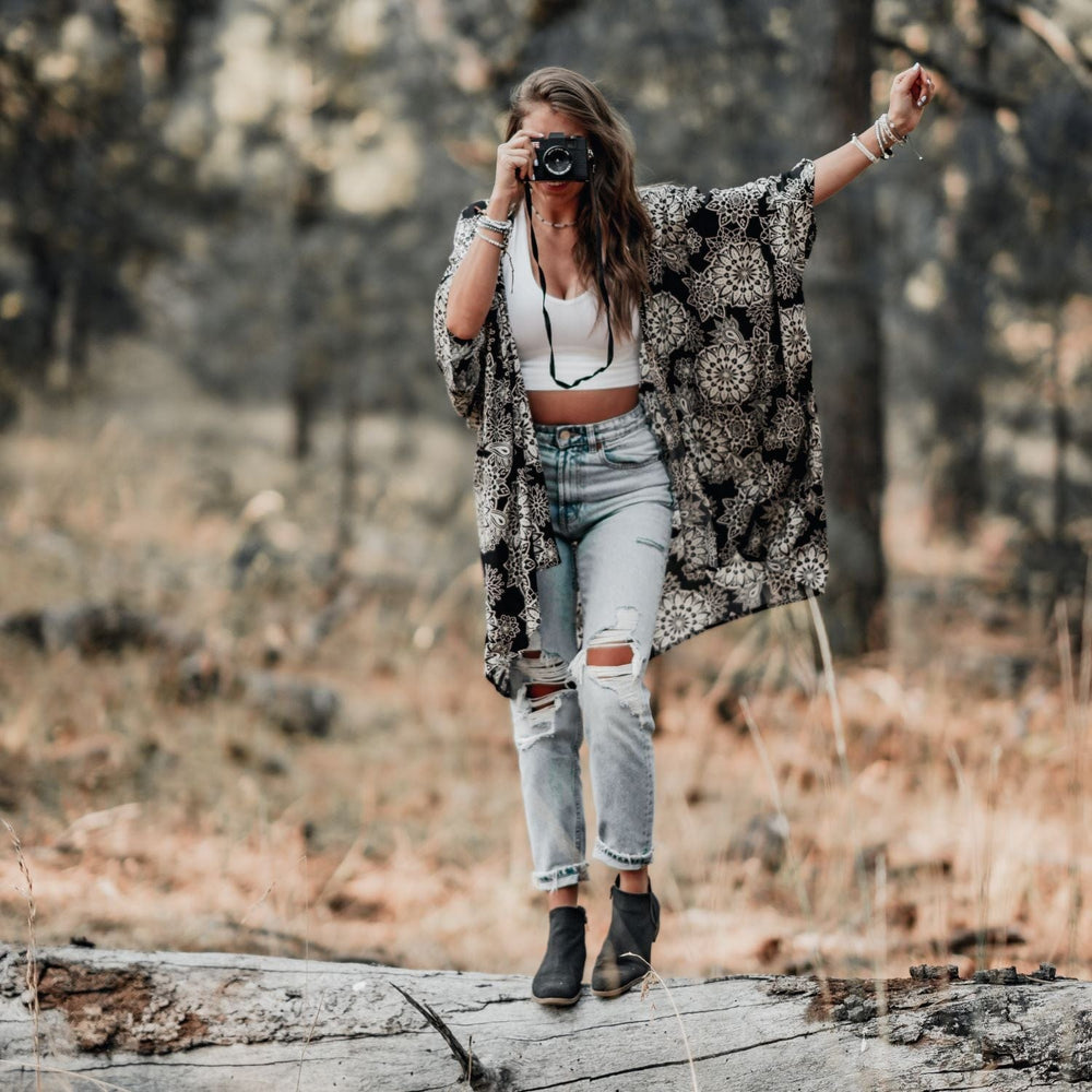 A woman taking a photo with a camera, standing on a fallen tree trunk, wearing torn jeans, white rop top, black and white kimono style coverup, black ankle boots, and wearing a stack of beaded bracelets on each wrist, with burred tan green woods in background.