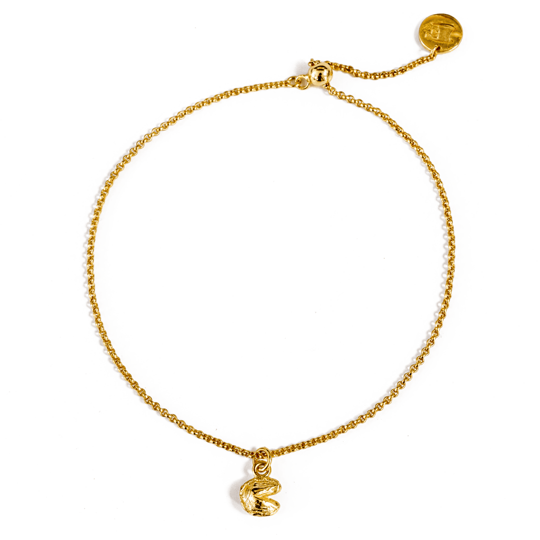 Close-up of a gold chain bracelet with a claw clasp closure, gold logo charm, and gold fortune cookie charm, displayed on a transparent background.