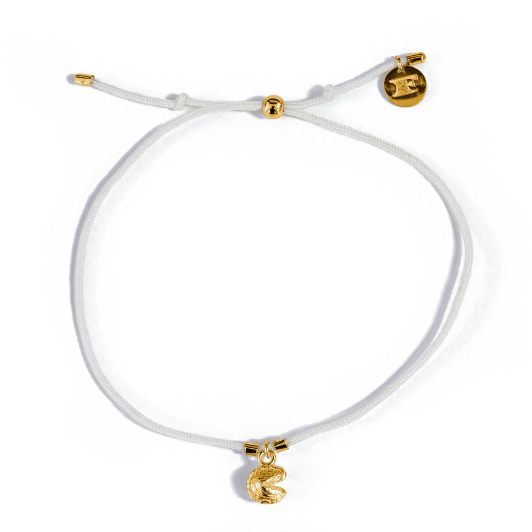 Close-up of a creamy white string bracelet with pull tie closure, gold accent beads and logo charm, and gold fortune cookie charm, displayed on a transparent background.