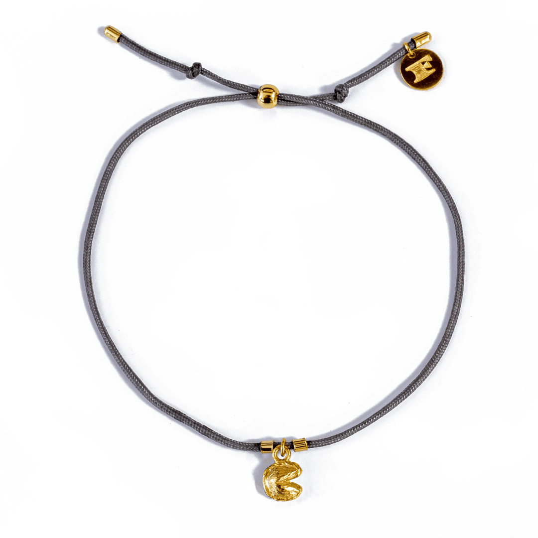 Close-up of a gray string bracelet with pull tie closure, gold accent beads and logo charm, and gold fortune cookie charm, displayed on a transparent background.