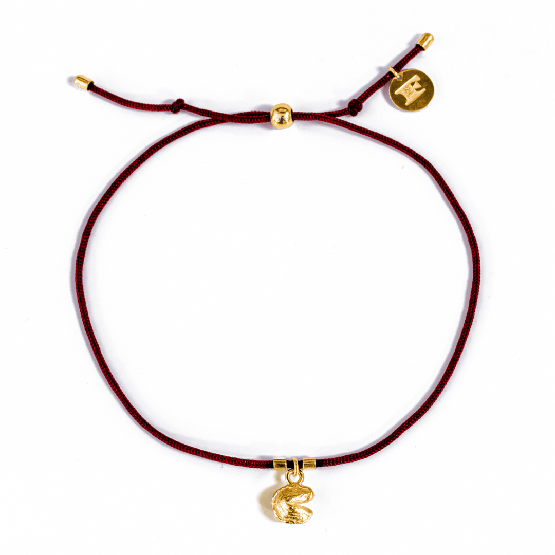 Close-up of a red string bracelet with pull tie closure, gold accent beads and logo charm, and gold fortune cookie charm, displayed on a transparent background.