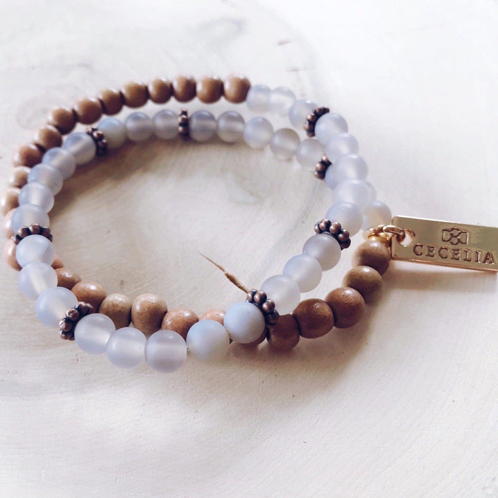 A stretch syle double-wrap bracelet made of frosted gray, and brown wood, beads with copper accent beads, and a gold-tone logo tag, displayed on a light tan wood background.