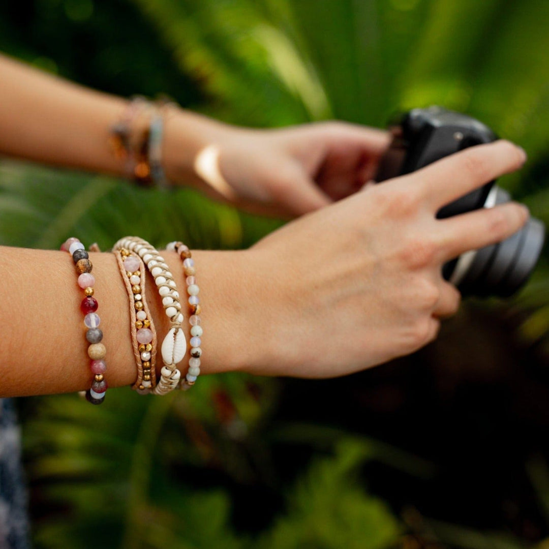 Close-up of a woman's hands holding a camera with multiple bead bracelets on her wrist, including the Hang Ten macrame bracelet, with blurry green palm fronds in the background.