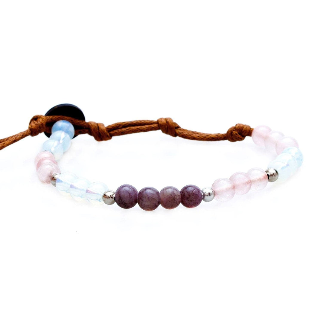 Close-up of a healing bead bracelet made with 6mm multicolor round moonstone and rose quartz beads, in shades of blue, purple, pink, and white colors, with silver accent beads, and a brown adjustable cord with a dark brown coconut shell button closure, displayed on a white background.