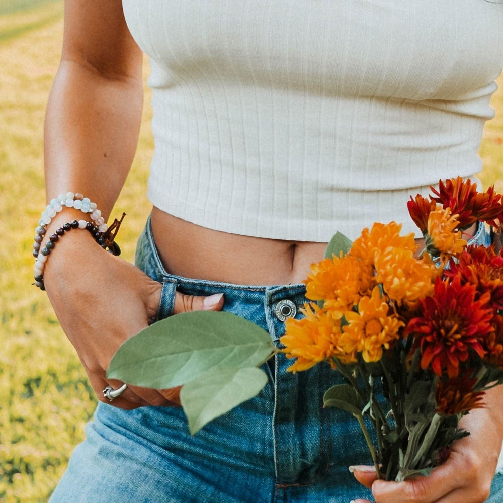 Close-up of a woman's torso wearing a white knit sleeveless crop top and blue jeans with her belly button showing, holding yellow and red flowers in one hand, and wearing three bracelets on her opposite wrist, including the healing and love bracelet, with blurred grass background.