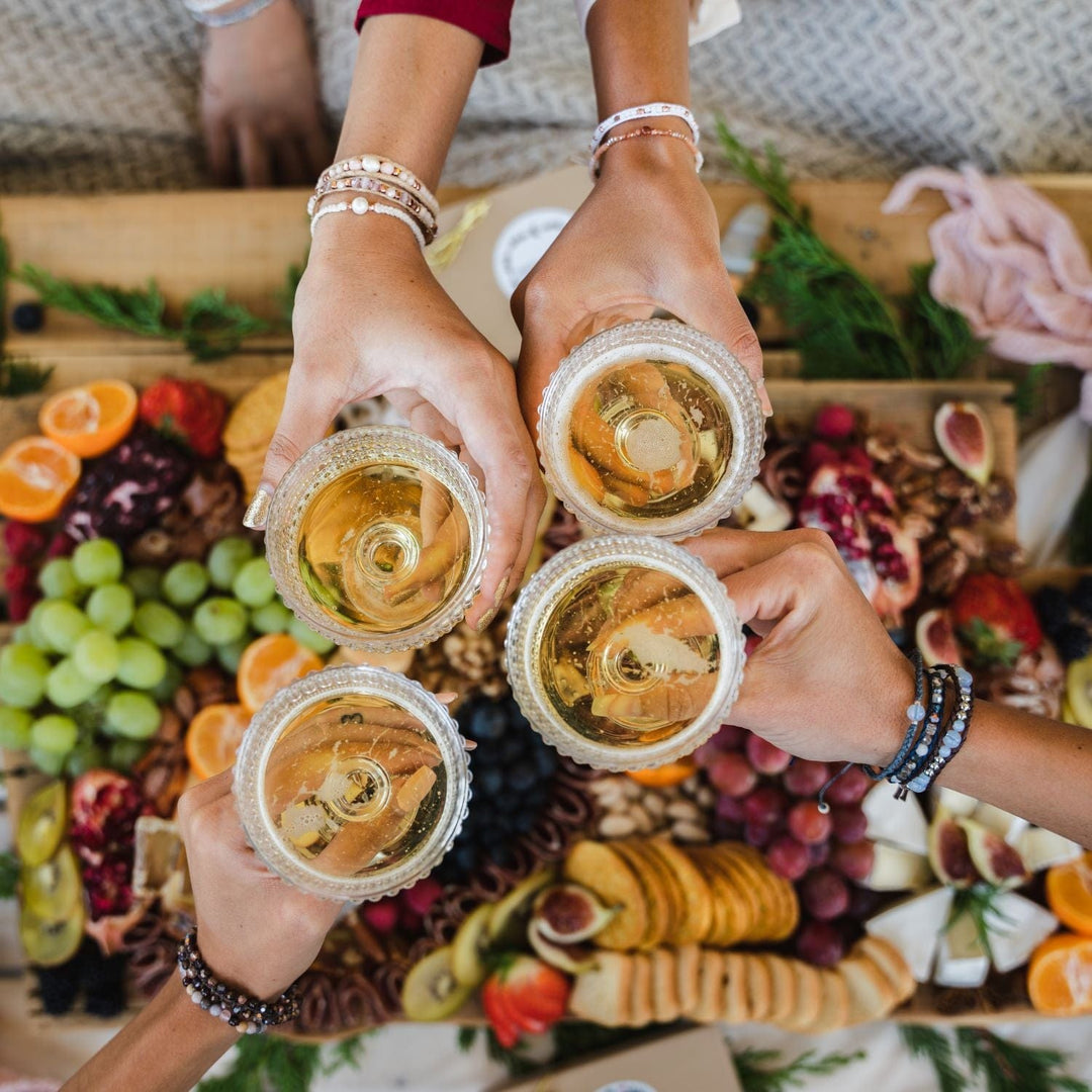 Close-up overhead view of four woman's hands toasting champagne on New Year's Eve, with a blurred charcuterie board in the background, each woman is wearing  beaded bracelets, one with three brcelets in blue shades, one with three bracelets in white and pink shades, one with two bracelets in white and pink colors, and one with three bracelets in brown and black colors.