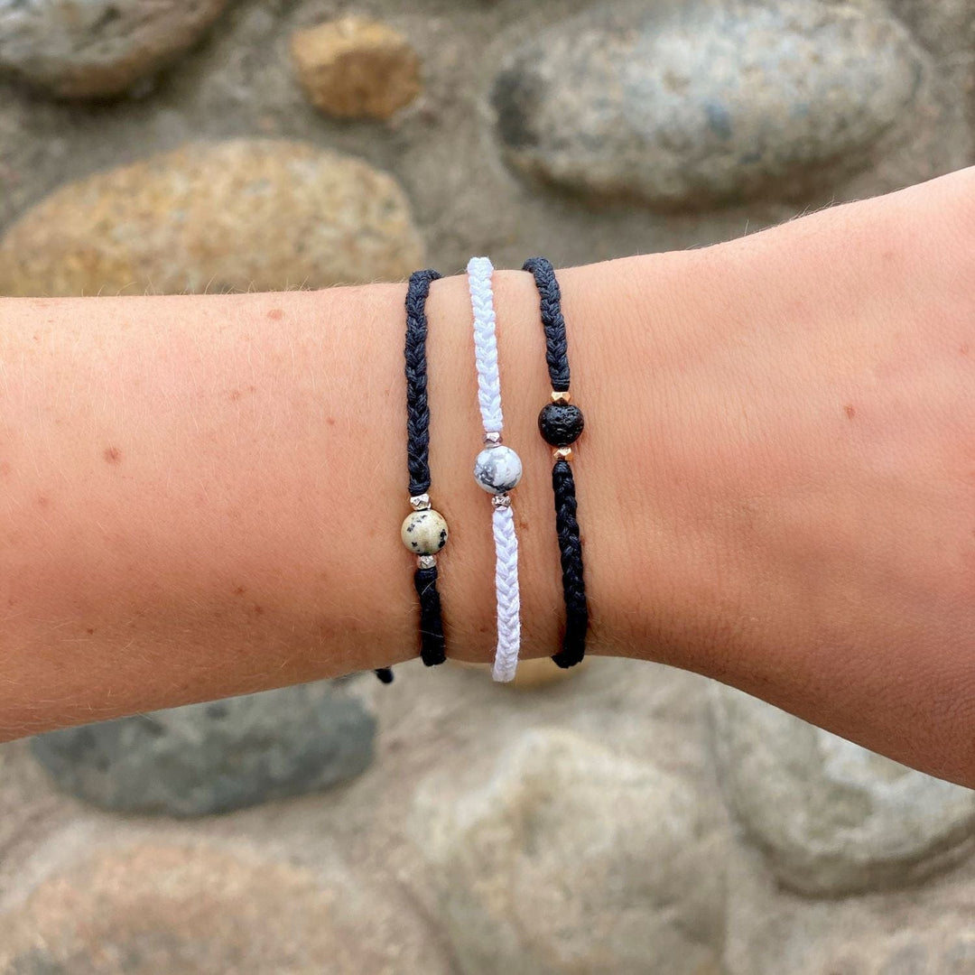 A close-up of a young woman's wrist wearing three braided bracelets with a center stone, one is black with a damatian asper stone, one is white with a white and gray howlite stone, and one is blacl with a black lava stone, with a rock wall in the background.