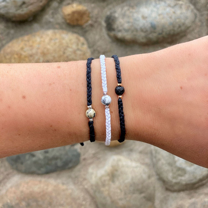 A close-up of a young woman's wrist wearing three braided bracelets with a center stone, one is black with a damatian asper stone, one is white with a white and gray howlite stone, and one is blacl with a black lava stone, with a rock wall in the background.